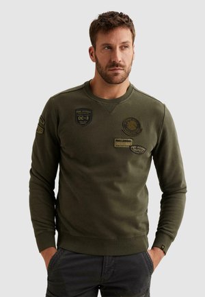 Mann trägt olivgrünes Sweatshirt mit militärisch inspirierten Abzeichen, Hände in den Taschen, schaut leicht zur Seite vor einfarbigem Hintergrund.