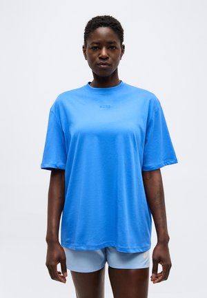 Svart kvinne iført en løs blå T-skjorte og lyseblå shorts står foran en ensfarget grå bakgrunn.