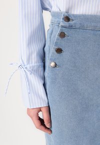 Jupe en denim bleu clair avec des boutons argentés sur le côté, associée à une chemise rayée bleu clair avec un détail de nœud au niveau des manches.