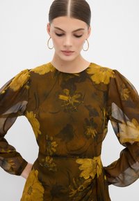 Bruine bloemenblouse met doorschijnende, gepofte mouwen. Bevat gele en donkere bloemenpatronen, een ronde halslijn en een getailleerde taille. Zachte textuur.