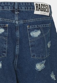 Shorts de mezclilla en un lavado azul medio con parches desgastados, bolsillos traseros y una etiqueta blanca marcada "RAGGED JEANS" en la cintura.