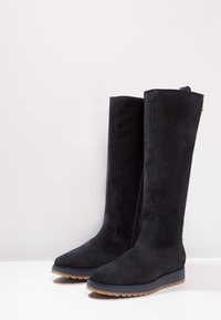 Botas altas de ante negras con suela plana, puntera redondeada y diseño minimalista. Con un lazo trasero para facilitar el uso.