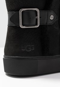 Botte en daim noir avec une semelle en caoutchouc, dotée d'une sangle à boucle métallique et du logo UGG embossé ; surface texturée avec une finition mate.