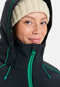 Veste extérieure noire avec des accents verts et un col haut, dotée d'une fermeture éclair et de cordons de serrage. Portée avec un bonnet tricoté de couleur claire.