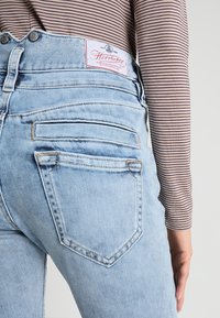 Světlé modré džíny s vysokým pasem, růžová nášivka, design s pěti kapsami a jemným prošíváním. Tkanina vypadá měkce a je mírně opotřebovaná.