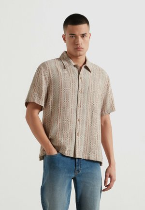 Mann mit kurzen Haaren, der ein beige gemustertes kurzärmeliges Hemd und blaue Jeans trägt, steht mit einer Hand in der Tasche vor weißem Hintergrund.