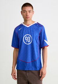 Zils futbola krekls, kas izgatavots no elpojoša auduma, ar šūnu rakstu, sarkaniem akcentiem un apli logotipā ar "90" centrā.