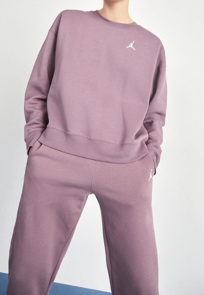 Personne portant un sweat-shirt et un pantalon de survêtement mauve assortis avec des logos Jumpman blancs, debout les mains dans les poches devant un fond uni.