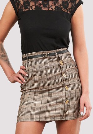 Kvinde iført sort blondetop og beige ternet knap-nederdel foran med sort bælte med dobbeltringspænde, poserer med hånden på hoften.