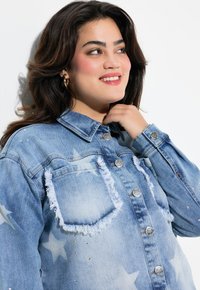 Chaqueta de mezclilla azul claro con patrones de estrellas, bordes deshilachados y acentos de rhinestones. Cuello clásico y cierres de botón plateados.