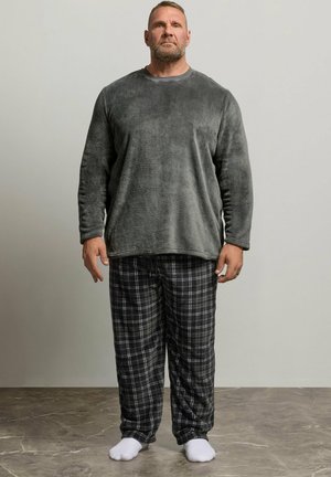 Homme debout pieds nus sur un sol carrelé portant un haut gris duveteux à manches longues et un pantalon à carreaux noir et blanc devant un mur uni.