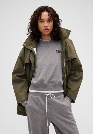 Femme aux cheveux bouclés portant une veste vert olive par-dessus un sweat-shirt gris GAP et un pantalon de jogging gris assorti, les mains dans les poches.