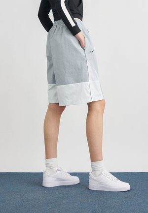 Osoba odjevena u svijetlosive i bijele duge kratke hlače, crnu majicu dugih rukava, bijele čarape i bijele Nike tenisice stoji na plavom tepihu.