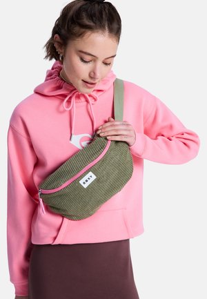 Joven con sudadera con capucha rosa y falda marrón sostiene una riñonera texturizada verde oliva con cremallera rosa sobre su pecho.