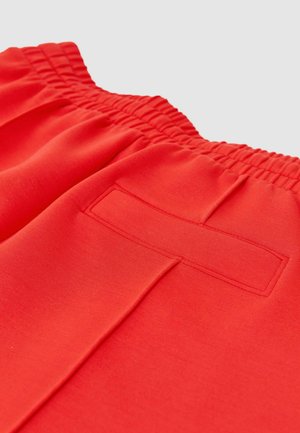 Pantalon à taille élastique rouge vif avec une poche passepoilée à l’arrière et un détail de couture centrale sur un fond clair uni.