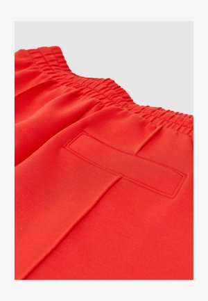 Pantalon à taille élastique rouge vif avec une poche passepoilée à l’arrière et un détail de couture centrale sur un fond clair uni.