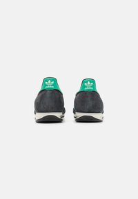 Tênis Adidas em camurça preta com detalhes em verde no calcanhar. Apresenta o logo icônico e sola em tom creme. A vista traseira mostra contornos suaves.
