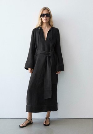 Frau trägt ein schwarzes, langärmliges, tailliertes Kleid, schwarze Sandalen und übergroße schwarze Sonnenbrillen, steht vor einer schlichten weißen Wand.