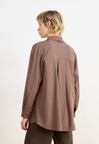 Camicia marrone dal taglio morbido con una leggera lucentezza, maniche lunghe, polsini abbottonati e un pannello posteriore plissettato. Presenta un colletto e un piccolo dettaglio ad anello.