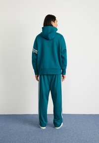 Felpa color teal con un grande cappuccio e strisce bianche su una manica, abbinata a pantaloni tuta teal con strisce bianche sui lati.