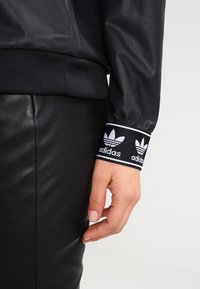 Czarna sportowa kurtka z prążkowanym mankietem z białym logo Adidas. Gładki materiał, dopasowany krój i kontrastujący pas w talii. Widoczna czarna skórzana spódnica.