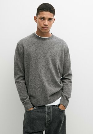 Junger Mann mit kurzen Haaren, der einen grauen Pullover und dunkle Jeans trägt, steht mit den Händen in den Taschen vor einem schlichten weißen Hintergrund.