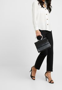 Borsa a mano in pelle nera con un design strutturato e un manico rotondo e trasparente, indossata con una camicia bianca con bottoni e pantaloni neri aderenti.