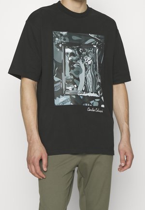 Person wearing black t-shirt with abstract grayscale face and frame graphic and "Carlo Colucci" text, paired with olive green pants.