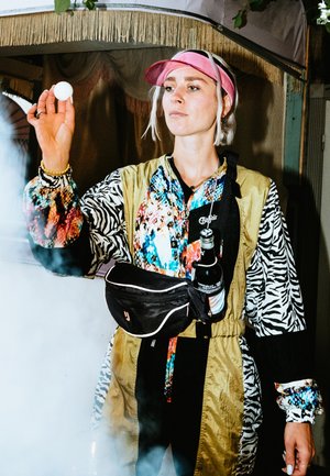 Colorful, patterned jacket featuring floral, zebra, and solid segments; equipped with a black waist bag and holding a ping pong ball.