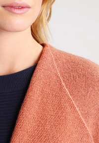 Světle broskvový, strukturovaný cardigan s překládaným designem, přeložený přes tmavě modrý žebrovaný svetr. Tkanina vypadá měkká a teplá.
