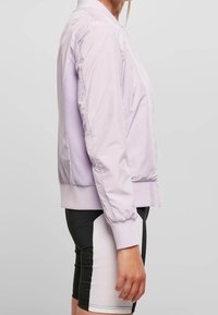 Jasnopurpurowa bomberka z teksturowanymi siateczkowymi bokami, elastycznymi mankietami i prążkowanym dołem. Noszona na czarno-białe szorty rowerowe.