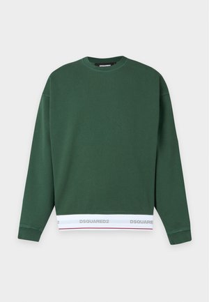 Jeune homme portant un sweat-shirt vert foncé, un jean skinny gris clair déchiré et des baskets noires, debout devant un fond blanc.