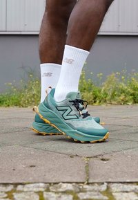 Sapatilhas New Balance de tom azul claro com detalhes em preto, tecido texturizado e uma sola grossa com detalhes em laranja, combinadas com meias brancas.