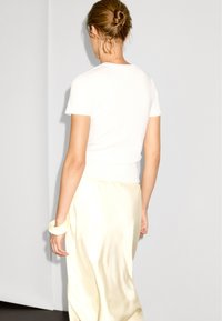 Bílý upnutý tričko s krátkými rukávy, spárované s saténovou krémovou sukní. Vlasy upravené do nízkého drdolu, minimální doplňky.