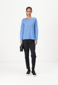 Ανοιχτό μπλε, χαλαρό πλεκτό πουλόβερ με φαρδύ λαιμό και ριγέ μα νσέτες, συνδυασμένο με μαύρο εφαρμοστό παντελόνι και μαύρα αθλητικά παπούτσια. Κρατά ένα μικρό μαύρο τσαντάκι.