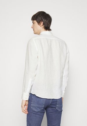 Camisa blanca de lino de manga larga con cuello abotonado, que presenta un dobladillo curvado y puños de un solo botón, combinada con jeans de denim azul.