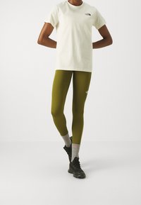 Osoba v bílém tričku s krátkým rukávem a olivově zelených legínách, s šedými ponožkami a černými teniskami na jednolitém pozadí.