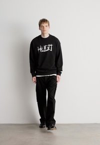 Μαύρη μπλούζα με λευκό γραφικό "HUGO", συνδυασμένη με μαύρα cargo παντελόνια και μαύρα παπούτσια. Το μοντέλο στέκεται απέναντι σε έναν απλό φόντο.