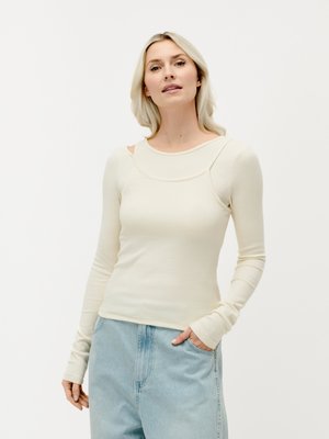 Blonde Frau trägt ein cremefarbenes Langarmoberteil mit Ausschnittdetails und hellblaue, hoch taillierte Jeans, steht mit einer Hand in der Tasche.