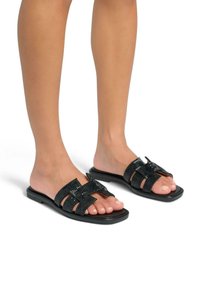 Chanclas negras con correas trenzadas, suelas planas y diseño de puntera abierta. Presentan un acabado brillante con un patrón texturizado en las correas.