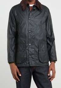 Tmavě zelená voskovaná bunda s hnědým manšestrovým límcem, druky a dvěma předními kapsami. Má texturovaný povrch a volný střih.