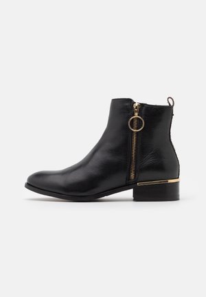 Bottine noire en cuir avec talon bloc bas, fermeture éclair dorée avec tirette ronde, et bordure métallique sur le bord du talon, vue de côté sur fond blanc.