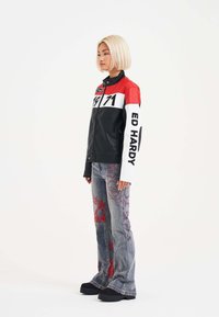 Chaqueta de cuero negra con acentos rojos y blancos, con cremalleras y etiqueta "ED HARDY". Jeans acampanados con patrones bordados en rojo. Zapatos de plataforma negros.