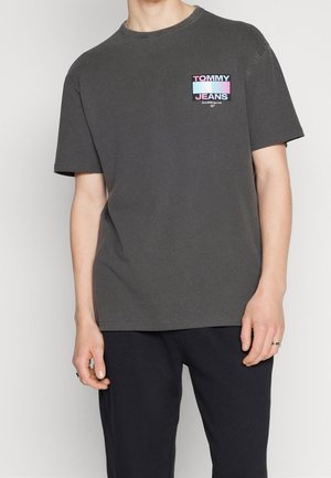 Mann trägt ein dunkelgraues Tommy Jeans T-Shirt mit kleinem pastellfarbenem Logo auf der Brust, kombiniert mit schwarzer Hose und Ringen an den Fingern.