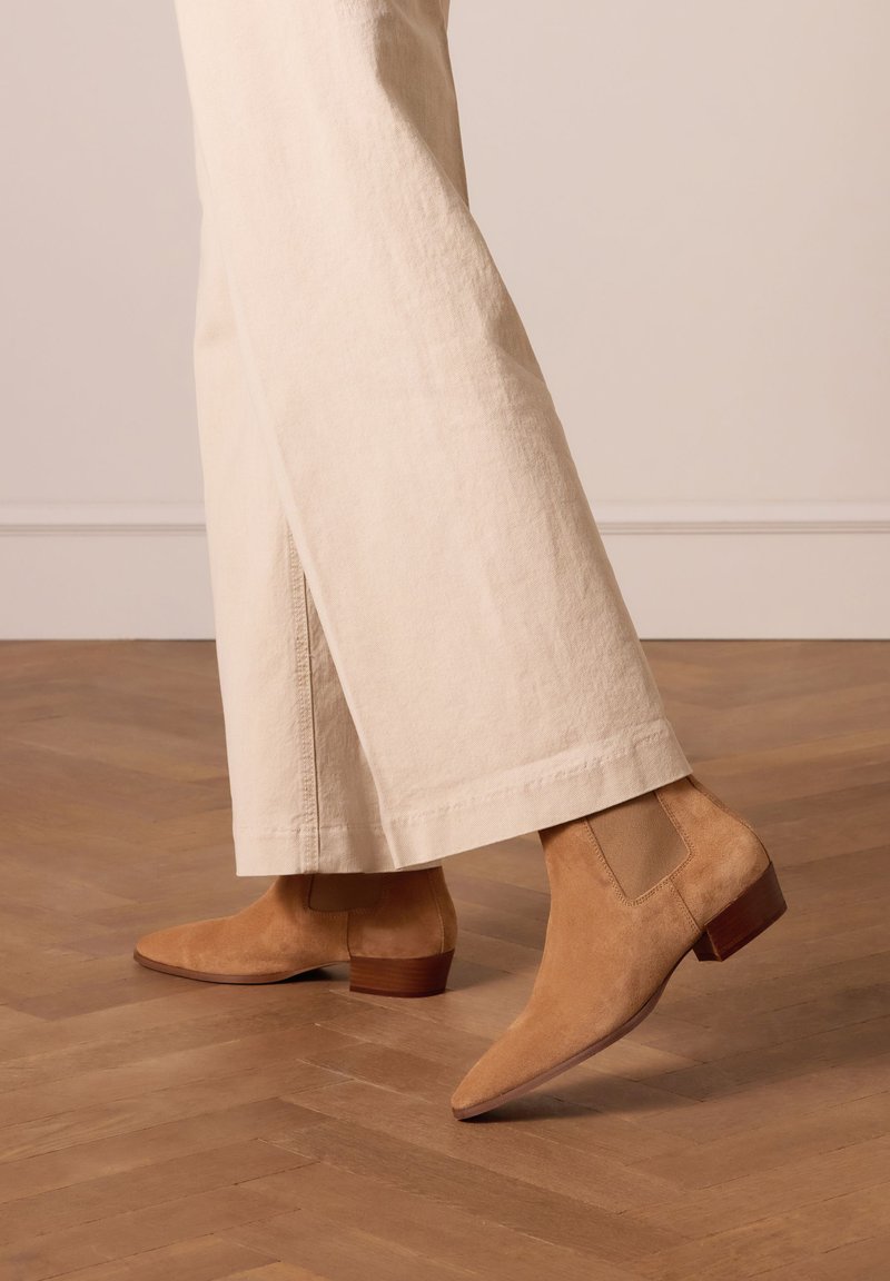 Tansuede enkellaarzen met een spitse neus en een lage blokhak, uitgerust met elastische zijpanelen voor eenvoudig aan- en uittrekken, gecombineerd met lichtbeige wijde broekspijpen.