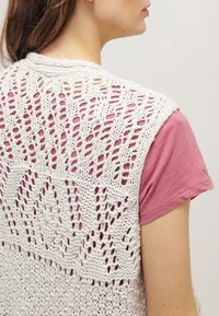 Pletený svetr v off-white barvě s otvorenými vzory; má texturovaný design s výrazným diamantovým motivem na zádech. V kombinaci s růžovou košilí.