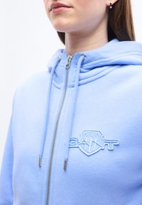 Γυναίκα που φοράει ανοιχτό μπλε φούτερ με φερμουάρ, κορδόνια και κεντητό λογότυπο GANT στο στήθος, που δείχνει το κάτω μέρος του προσώπου και τον λαιμό.