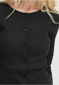 Černý žebrovaný kardigan s kulatým výstřihem, vybavený šesti knoflíky potaženými látkou a texturovaným designem. Měkký, přiléhavý materiál.