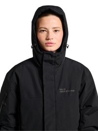 Nuori henkilö pukeutuneena mustaan hupulliseen takkiin, jossa on vetoketju ja nyöritykset, katsoo eteenpäin, neutraali ilme, teksti "HALO 196819721981" rinnassa.