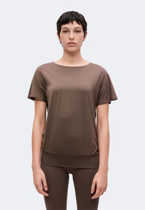 Mujer de cabello corto y oscuro que lleva una camiseta marrón de manga corta y pantalones a juego, de pie frente a un fondo liso y claro.